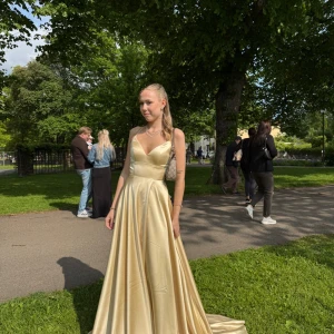 Dressroom linne klänning gul  - Originalpris 5500 kr. Säljer en elegant långklänning i champagnefärgad satin med smala axelband och öppen rygg med snörning. Klänningen har v-ringad urringning och ett svepande, glansigt tyg som ger en lyxig känsla. Perfekt för bal eller student!