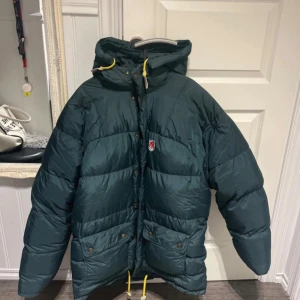 Mörkgrön pufferjacka från Fjällräven - Den är M men skulle säga att den är mer som L eller xl.  Enda slitaget är på drag kedjan där färgen har lossnat