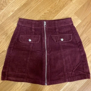 Vinröd manchesterkjol från Bershka - Vinröd kortkjol i manchester från Bershka med markerade fickor framtill, bälteshällor och en snygg dragkedja hela vägen fram. Kjolen har en cool retrovibe och passar perfekt till stickade tröjor eller t-shirts.