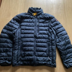 Svart dunjacka Parajumpers - Snygg svart dunjacka från Parajumpers i super lättviktsmodell. Jackan har quiltad design, hög krage med gul detalj, flera dragkedjor och patch på ärmen. Perfekt för dig som vill ha en stilren och varm jacka med coola detaljer.