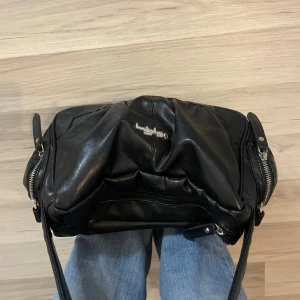 Svart axelväska från L.Credi i skinn - Cool svart axelväska från L.Credi i mjukt skinn med silverfärgad logga framtill. 🌸Väskan har dragkedjor på sidorna och en bred axelrem som gör den enkel att bära. Perfekt storlek för att få plats med allt du behöver och enkel att styla 💗🙌🏼