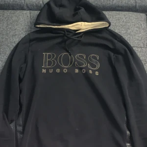 Svart hoodie från Hugo Boss - Svart hoodie från Hugo Boss med stor BOSS-logga framtill i beige. Tröjan har huva med beige insida och snörning. Perfekt för dig som gillar stilrena och klassiska streetwear-plagg. Långärmad och i mjukt material.