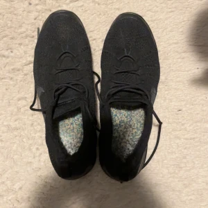 Nike Vapormax Flyknit svarta sneakers  - Svarta Nike Vapormax Flyknit sneakers med stickad ovandel och snörning. Snygg, rund tå och platt sula med synlig luftdämpning. Diskret Nike-logga på sidan och Vapormax-märke på plösen. Perfekt för dig som gillar sportig och modern stil.