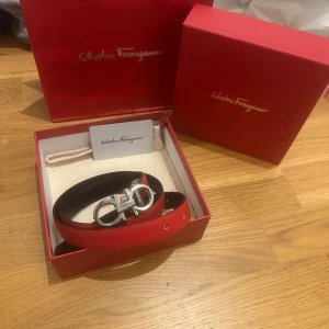 Ferragamo bälte - Snyggt rött bälte från Salvatore Ferragamo i skinn med ikoniskt silverfärgat spänne. Bältet har en stilren design med svarta kanter och klassisk Ferragamo-logga på spännet. Perfekt accessoar för att lyfta din outfit. Lite slimmare modell för women men funkar till alla