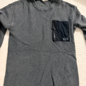 Grå stickad tröja från H&M med ficka - Grå stickad tröja från H&M med lång ärm och cool svart bröstficka med dragkedja och tryck. Tröjan har ett rutigt stickat mönster och rund halsringning. Perfekt för dig som gillar en avslappnad men ändå stilren look.