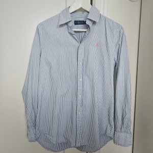 Randig skjorta från Ralph Lauren - Klassisk skjorta från Ralph Lauren i blåvitt randigt mönster. Skjortan har långa ärmar, knappar framtill och ikonisk rosa broderad logga på bröstet. Tillverkad i mjuk bomull och har en tidlös, avslappnad passform.