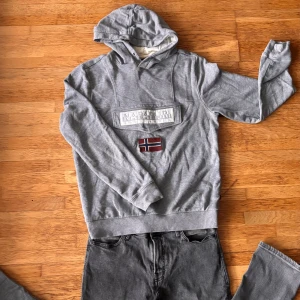 Grå hoodie från Napapijri med flagga - Säljer en grå hoodie från Napapijri med broderad norsk flagga på bröstet och stor logga framtill. Tröjan har huva, långa ärmar och är tillverkad i mjuk bomull. Perfekt för dig som gillar streetwear och snygga detaljer.