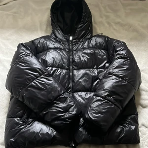 Svart pufferjacka med luva  - Säljer en svart glansig pufferjacka i storlek M. Jackan har en stor huva, dragkedja framtill och två sidofickor. Det har som man ser på sista bilden en liten skråma. 