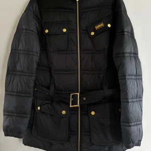 Svart pufferjacka från Barbour - Svart pufferjacka från Barbour med guldfärgade knappar och detaljer. Jackan har hög krage, bälte i midjan med metallspänne, flera fickor med lock och dragkedja framtill. Perfekt för kalla dagar och har en cool, urban vibe.