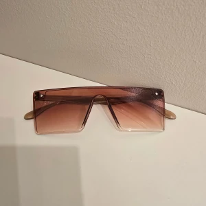 Solglasögon - Säljer ett par trendiga oversized solglasögon med rektangulära, beige tonade linser och transparent båge. Glasögonen har en futuristisk vibe och passar perfekt för dig som vill sticka ut med din stil.