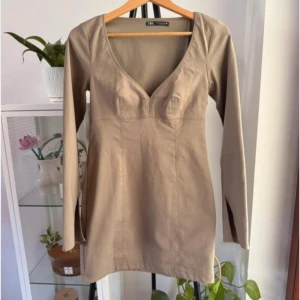 Beige kort klänning från Zara - Supersnygg beige kort klänning från Zara med v-ringad hals och långa ärmar med slits. Figurnära passform som framhäver siluetten. Perfekt för dig som gillar minimalistisk stil och vill ha något som sticker ut med coola detaljer. Klänningen är strl L, bilderna 2 & 3 visar hur de sitter på men kan vara en annan storlek 