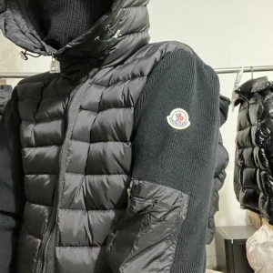 Svart Moncler liknande pufferjacka med huva - Alla storlekar finns inne. Skriv privat, leverans tar runt 2 veckor  Svart pufferjacka som liknar Moncler med stickade ärmar och sidopaneler, huva med snörning och dragkedja framtill. Jackan har en quiltad front och ärm med ficka och en Moncler liknande logga på vänster arm. Materialmix av polyester och stickat ger en schysst look.