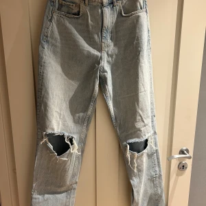 Ljusblå ripped jeans från Gina Tricot - Ljusblå jeans från Gina Tricot med snygga slitningar och stora hål vid knäna. Klassisk femficksmodell med hög midja och raka ben. Jeansen är i denim och har en avslappnad vibe, perfekt för dig som gillar en trendig och edgy stil.