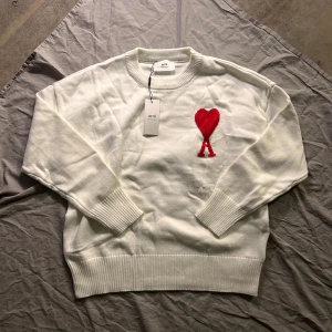 Vit stickad tröja med rött hjärta från Ami - Vit stickad tröja från Ami med rund hals och ribbade muddar. På bröstet finns ett stort rött hjärta med bokstaven A under, som är typiskt för Ami de Coeur. Tröjan har lång ärm och är gjord i mjuk bomull, perfekt för att ge din outfit en clean vibe.
