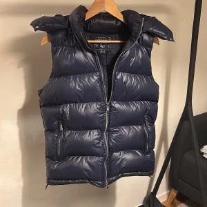 Mörkblå dunväst från Leon Marcel - Snygg mörkblå dunväst från Leon Marcel med hög krage och avtagbar huva. Västen är helt o använd/ny och har två dragkedjefickor framtill och quiltad design som ger en puffig look. Perfekt för lager-på-lager och håller dig varm under kyliga dagar.