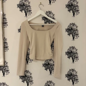 Beige långärmad topp från SHEIN - Snygg beige långärmad topp från SHEIN med bred, fyrkantig urringning. Modellen är croppad och har en stilren, slät design utan mönster eller detaljer. Perfekt för dig som gillar minimalistisk stil och vill ha en enkel topp att matcha med allt.