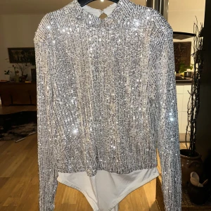 Glittrig silver bodysuit med paljetter - Säljer en glittrig bodysuit i silver med långärmad design och hög hals. Toppen är täckt av paljetter som ger ett riktigt festligt intryck och har en slät, vit underdel. Perfekt för dig som vill sticka ut på nästa event!