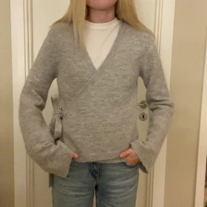Grå omlottkofta med knyt i sidan - Mysig grå stickad omlottkofta med v-ringning och knytband i sidan. Är i mycket bra skick