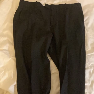 Svarta kostymbyxor från Steel - Snygga svarta kostymbyxor från Steel med klassisk rak passform och diskret knapp- och dragkedjestängning. Byxorna har bälteshällor och är tillverkade i ett mjukt material som ger en stilren look. Perfekta för dig som gillar en clean och enkel stil.