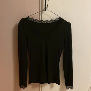 Svart långärmad topp med spetsdetaljer - (Den är oanvänd) Säljer en svart långärmad topp med snygga spetsdetaljer vid halsringningen och ärmsluten. Toppen har en figurnära passform och är perfekt för dig som gillar en stilren look med en touch av romantik. Materialet känns mjukt och stretchigt.💕