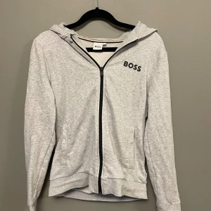 Grå hoodie från Hugo Boss med dragkedja - Grå hoodie från Hugo Boss med svart logga på bröstet. Tröjan har huva, hel dragkedja framtill och två fickor. Tillverkad i mjuk bomullsmix, perfekt för chill dagar. Långärmad och med ribbade muddar för extra komfort.