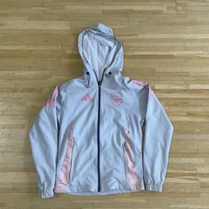 Vit vindjacka Arsenal x Adidas - Snygg vit vindjacka från Adidas i samarbete med Arsenal, med rosa detaljer och logga på bröstet. Jackan har huva med snörning, hel dragkedja och långa ärmar. Perfekt för dig som gillar sportig stil och vill sticka ut med coola färgdetaljer.