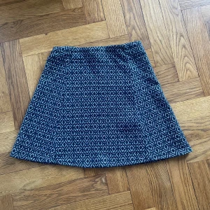 Mönstrad blå kortkjol från Zara - Snygg kortkjol från Zara med geometriskt mönster i mörkblått och vitt. Kjolen har en A-linjeformad siluett och är tillverkad i ett mjukt syntetmaterial som känns bekvämt mot huden. Perfekt för dig som gillar coola mönster och vill sticka ut lite extra.