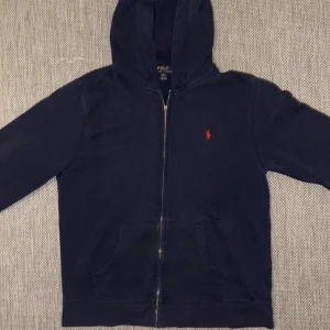 Marinblå hoodie från Polo Ralph Lauren - Snygg marinblå hoodie från Polo Ralph Lauren med dragkedja och klassisk röd logga på bröstet. Tröjan har huva, känguruficka och långa ärmar. Perfekt för en chill och avslappnad stil.