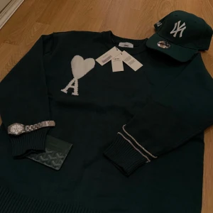 Stickat tröja mörkgrön Ami - Snygg mörkgrön stickad tröja från Ami med vit hjärtlogga och bokstaven A på bröstet. Tröjan har rund halsringning, ribbade muddar och en clean look som passar perfekt till jeans. Materialet är mjukt och skönt, perfekt för kyligare dagar.