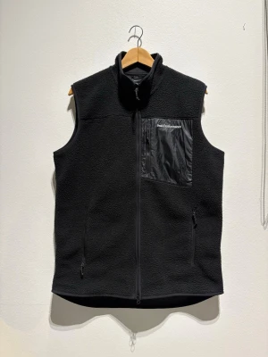 Svart fleeceväst Peak Performance L - Svart fleeceväst från Peak Performance i storlek L. Västen har en hel dragkedja framtill och en bröstficka i blankt material med dragkedja. Två sidofickor med dragkedja. Perfekt lager-på-lager-plagg för kyliga dagar. Aldrig använd 