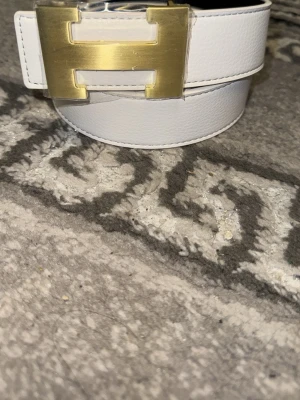 Vit skärp med H- guldspänne - Säljer ett stilrent vitt skärp med ett stort, fyrkantigt guldspänne. Skärpet är i slätt material och har en clean look som passar till många outfits. Perfekt accessoar för att lyfta din stil.