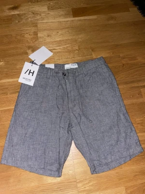 Grå shorts från Selected Homme - Säljer ett par grå shorts från Selected Homme i mjuk bomull med dragsko i midjan och knappgylf. De har en avslappnad passform, sidofickor och bakfickor med knapp. Perfekta för sommaren och tillverkade av ekologisk bomull. Helt oanvända! 