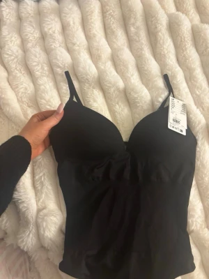 Svart body med tunna axelband - Säljer en svart body med tunna, justerbara axelband och vadderade kupor. Bodyn har en enkel och stilren design i mjukt material som sitter tight mot kroppen. Perfekt att styla under en skjorta eller kavaj för en clean look.