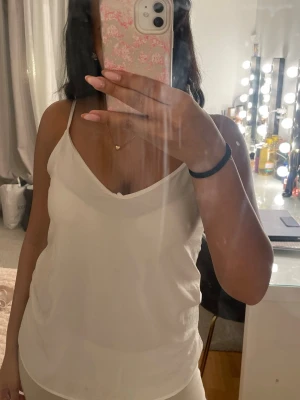 Vit linnetopp med tunna axelband - Säljer en enkel och stilren vit linnetopp med tunna axelband och v-ringad hals. Toppen har en lös passform och är tillverkad i ett lätt, luftigt material som känns skönt mot huden. Perfekt för varma dagar eller att styla lager på lager.