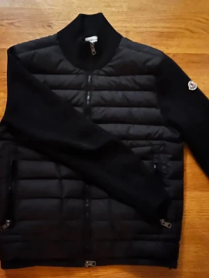 Moncler cardigan - Säljer en svart cardigan från Moncler med quiltad front och släta stickade ärmar. Jackan har dragkedja framtill, två sidofickor med dragkedja och Moncler-logga på ärmen. Perfekt för dig som vill ha en clean och stilren look.