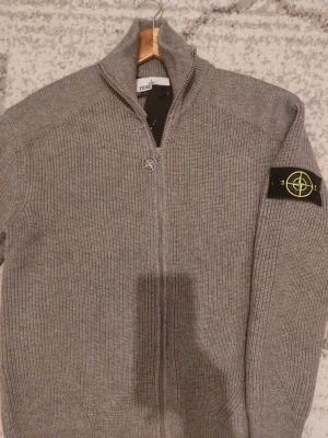 Grå stickad kofta från Stone Island - Säljer en grå ribbstickad kofta från Stone Island med dragkedja framtill och ikonisk patch på ärmen. Tröjan har hög krage och långa ärmar, perfekt för kyliga dagar. Klassisk och stilren design som funkar till det mesta. En extra badge läggs till på köpet.
