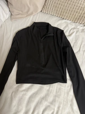 Svart croppad långärmad polotopp - Säljer en svart croppad polotopp med lång ärm och half zip-dragkedja framtill. Toppen är ribbad och har en tight passform som sitter snyggt på kroppen. Perfekt till jeans eller kjol för en clean look.