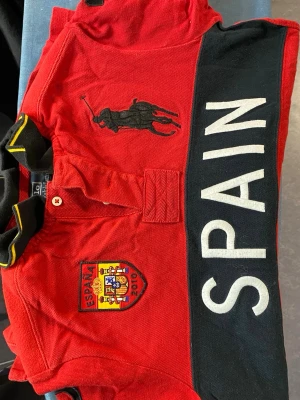 Röd Polo Ralph Lauren Spanien piké - Säljer en röd pikétröja från Polo Ralph Lauren med svart krage och gula detaljer. Stor broderad logga, Spanien-emblem och texten SPAIN framtill. Nummer 10 på ryggen och ärmen samt spansk flagga. Tillverkad i bomull och polyester, custom fit.