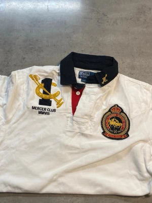 Vit pikétröja Polo Ralph Lauren S - Säljer en vit pikétröja från Polo Ralph Lauren i custom fit med svart krage och röda detaljer vid knäppningen. Tröjan har stora broderade märken på bröstet och är kortärmad. Klassisk och sportig look med Mercer Club och Polo Team-emblem.