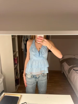 Ljusblå volangig halterneck  - Supersöt ljusblå linnetopp med volanger och v-ringning. Toppen är ärmlös och har knyt i nacken samt smockad rygg för extra komfort. Perfekt för varma dagar och ger en romantisk vibe till outfiten.