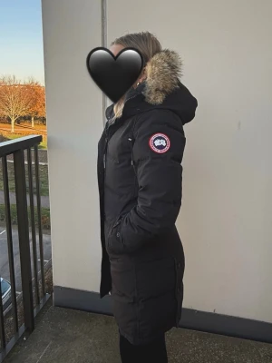 Svart dunjacka från Canada Goose - Säljer en svart lång dunjacka från Canada Goose med pälskantad huva och klassisk logga på ärmen. Jackan har dragkedja och knappar framtill, samt praktiska fickor och justerbar huva. Vid snapp affär kan jag släppa den för 3700 