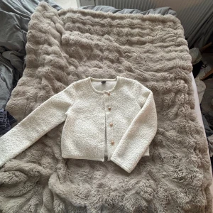 Vit kofta från Gina Tricot med guldknappar - Säljer en vit kofta från Gina Tricot i storlek M. Koftan har en fluffig, strukturerad yta och stängs med dekorativa guldknappar framtill. Rund halsringning och långärmad modell som passar perfekt till lager på lager under höst och vinter.