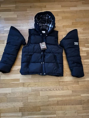 Svart dunjacka från Burberry med avtagbara armar - Snygg svart dunjacka från Burberry med avtagbara ärmar och huva. Jackan har dragkedja och knappar framtill, två fickor med dragkedja och klassiskt rutigt foder i huvan. Helt ny, aldrig använd, lappen sitter kvar. Storlek medium
