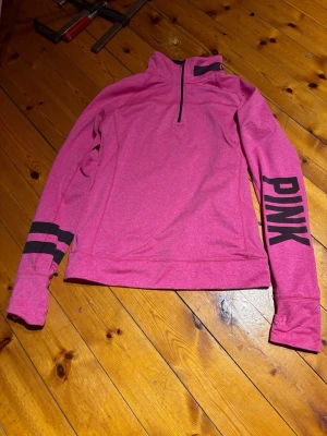 Rosa half zip tröja från PINK - Sportig rosa tröja från PINK by Victoria's Secret med svart tryck längs ena ärmen och två svarta ränder på andra ärmen. Tröjan har half zip, hög krage och är tillverkad i mjuk polyesterblandning. Perfekt för chill eller träning.