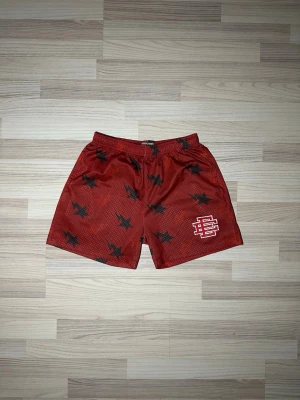 Röda meshshorts med stjärnor EE - Säljer ett par röda meshshorts från EE med svarta stjärnor över hela plagget och vit logga på ena benet. Shortsen har elastisk midja med vit snörning och är tillverkade i luftig polyester, perfekta för sommardagar eller träning. Liten passform 