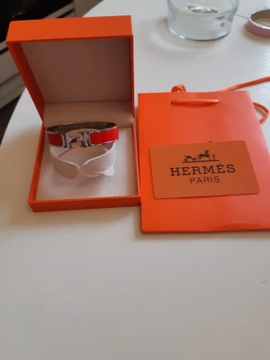 Hermes armband  - Bilderna visar ett Hermès-armband i röd och silverfärg med tillhörande ask och påse, 