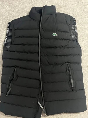 Svart dunväst från Lacoste - Säljer en svart dunväst från Lacoste med broderad krokodil på bröstet och diskreta Lacoste-loggor vid ärmöppningarna. Västen har två fickor med dragkedja och quiltad design. Perfekt för lager-på-lager och riktigt snygg till streetwear.