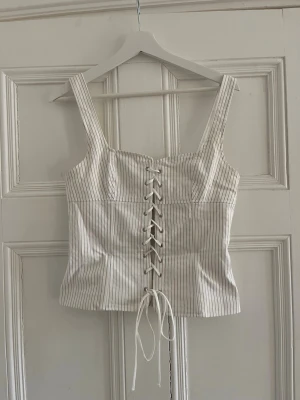 Trendig randig korsettopp från Zara - Snygg vit korsettopp från Zara med tunna svarta ränder och snörning framtill. Toppen har breda axelband och croppad passform.  Tveka inte på att höra av dig om du har frågor❤️