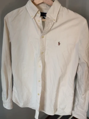 Vit skjorta från Ralph Lauren M - Klassisk vit skjorta från Ralph Lauren i custom fit. Skjortan har lång ärm, knappar framtill och ikonisk broderad logga på bröstet. Tillverkad i bomull och passar perfekt till en stilren look.