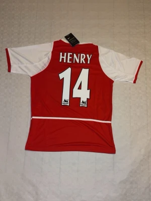 Arsenal Nike tröja Henry 14 röd/vit - Arsenal matchtröja från Nike med Henry 14-tryck på ryggen. Röd kropp med vita ärmar och detaljer, broderat Arsenal-märke och O2-logga framtill. Tillverkad i lätt polyester med Dri-FIT-teknik för skön känsla. Perfekt för fotbollsfans!